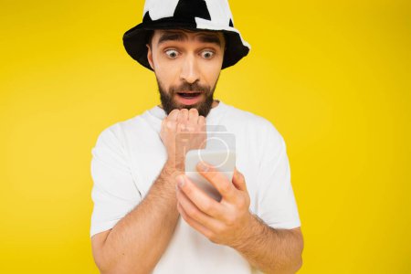 nervous man in football fan hat watching championship on smartphone isolated on yellow