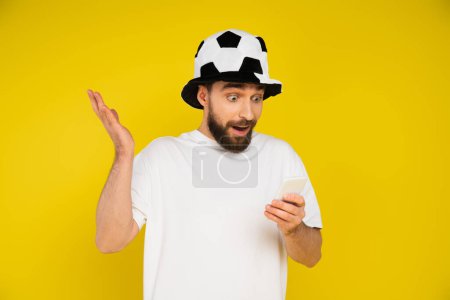 Foto de Sorprendido ventilador de fútbol en sombrero viendo juego en el teléfono móvil aislado en amarillo - Imagen libre de derechos