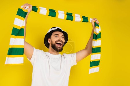 Foto de Abanico deportivo lleno de alegría en el sombrero gritando mientras sostiene la bufanda a rayas en las manos levantadas aisladas en amarillo - Imagen libre de derechos