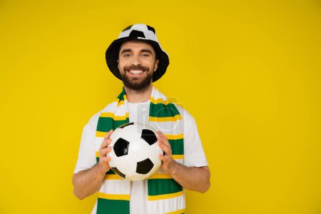 Foto de Hombre feliz en el sombrero del ventilador del fútbol y bufanda rayada sosteniendo pelota de fútbol aislado en amarillo - Imagen libre de derechos
