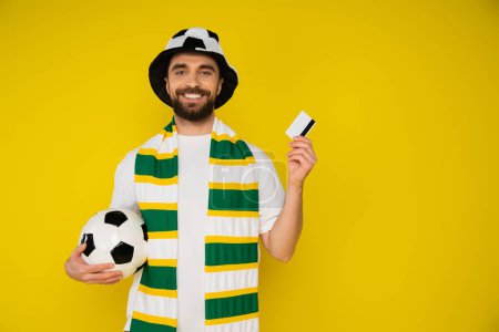cheerful football fan with soccer ball and credit card looking at camera isolated on yellow