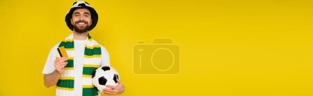 happy man in football fan hat standing with credit card and soccer ball isolated on yellow, banner