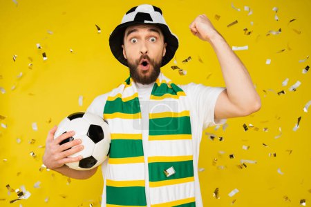 Foto de Admirador de fútbol sorprendido en sombrero y bufanda que muestra gesto de triunfo bajo la caída de confeti sobre fondo amarillo - Imagen libre de derechos