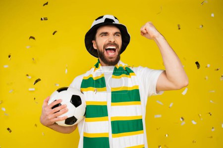 thrilled sports fan with soccer ball shouting and showing win gesture under falling confetti on yellow background