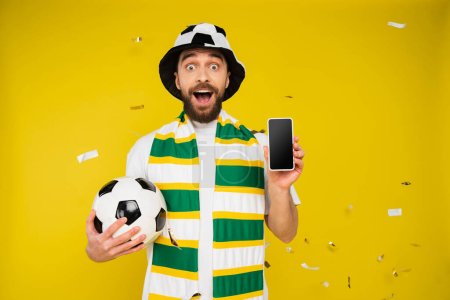 erstaunter Sportfan in gestreiftem Schal mit Fußballball und Smartphone mit leerem Bildschirm auf gelb 