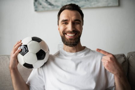happy bearded man looking at camera and pointing at soccer ball at home