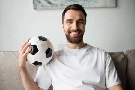 alegre y barbudo ventilador de fútbol sosteniendo pelota de fútbol y sonriendo a la cámara