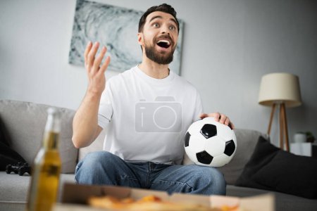 Photo for KYIV, UKRAINE - OCTOBER 21, 2022: cheerful man holding football and gesturing while watching championship at home - Royalty Free Image