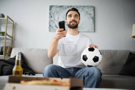 Photo for KYIV, UKRAINE - OCTOBER 21, 2022: cheerful man holding football and remote controller while watching championship at home - Royalty Free Image