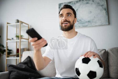 hombre feliz sosteniendo el fútbol y el mando a distancia mientras ve el campeonato en casa