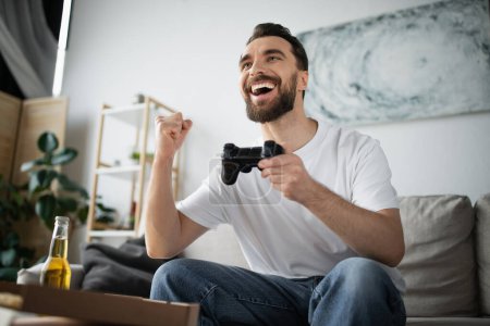 Foto de KYIV, UCRANIA - 21 de octubre de 2022: hombre emocionado jugando un videojuego cerca de la pizza y la botella de cerveza en casa - Imagen libre de derechos