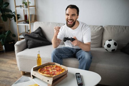 Foto de KYIV, UCRANIA - 21 de octubre de 2022: hombre barbudo feliz jugando videojuego cerca de la pizza y la botella de cerveza en casa - Imagen libre de derechos