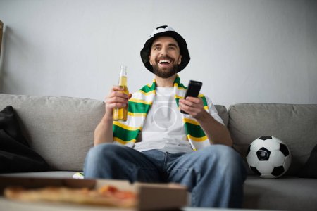 Photo for Happy man in fan hat and scarf holding remote controller and bottle of beer while watching football game - Royalty Free Image
