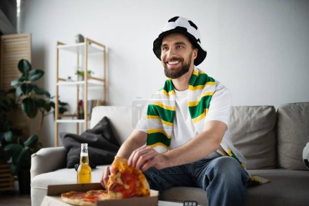 joyful man in hat and scarf taking slice of tasty pizza while watching championship