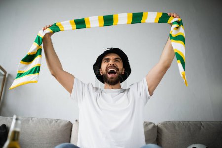 Foto de Hombre excitado en sombrero de fan sosteniendo bufanda estirado mientras grita y viendo el campeonato - Imagen libre de derechos