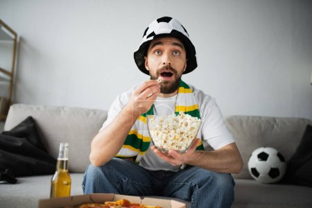 Foto de KYIV, UCRANIA - 21 de octubre de 2022: sorprendido hombre con sombrero deportivo y bufanda comiendo palomitas de maíz y viendo el campeonato - Imagen libre de derechos