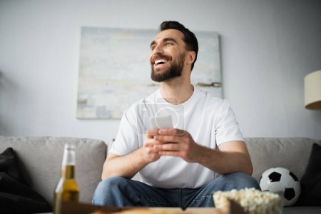 cheerful man using smartphone near bottle of beer and tasty food on blurred foreground 