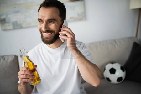 Foto de Hombre alegre sosteniendo la botella de cerveza y hablando en el teléfono inteligente cerca del fútbol en el sofá borroso - Imagen libre de derechos