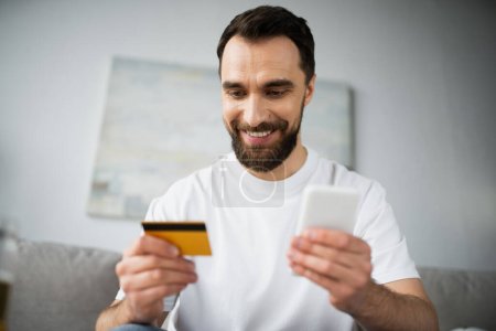 Photo pour Homme joyeux tenant carte de crédit et en utilisant smartphone tout en faisant des achats en ligne à la maison - image libre de droit