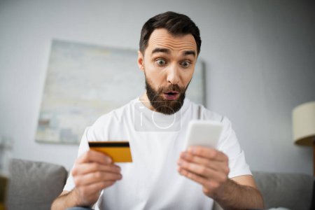 Photo pour Homme choqué tenant carte de crédit et en utilisant un smartphone tout en faisant des achats en ligne à la maison - image libre de droit