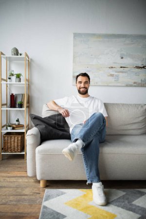 glücklicher bärtiger Mann in Jeans sitzt auf Couch im modernen Wohnzimmer 