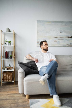 bärtiger Mann in Jeans sitzt auf Couch im modernen Wohnzimmer 