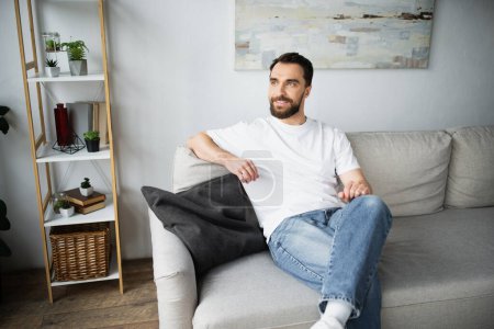 zufriedener und bärtiger Mann in Jeans sitzt auf Couch im modernen Wohnzimmer 