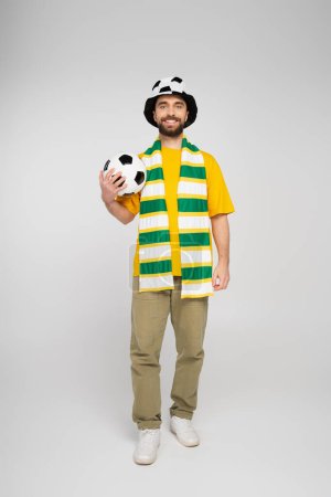 full length of happy bearded sports fan in hat holding football on grey