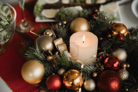 Tannenkranz mit Weihnachtskugeln und brennender Kerze auf Tisch mit festlichem Abendessen auf verschwommenem Hintergrund
