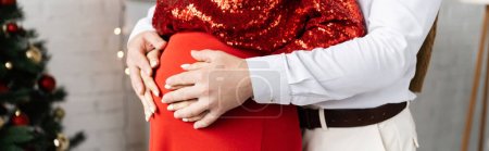 partial view of man embracing belly of pregnant woman in festive clothes on Christmas day, banner