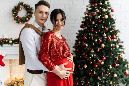 Schwangere und glückliche Asiatin mit Ehemann, der ihren Bauch in der Nähe des geschmückten Weihnachtsbaums umarmt