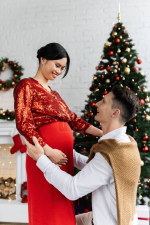 young man and his pregnant asian wife in festive clothing smiling at each other near blurred Christmas tree