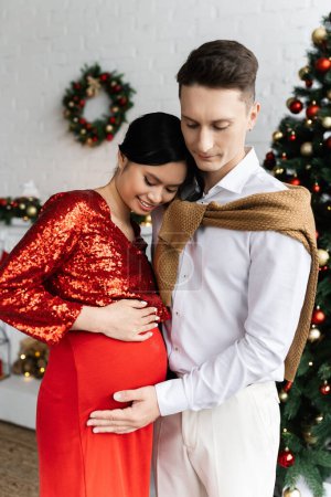 man touching tummy of smiling and pregnant asian wife in festive clothing near blurred Christmas decor
