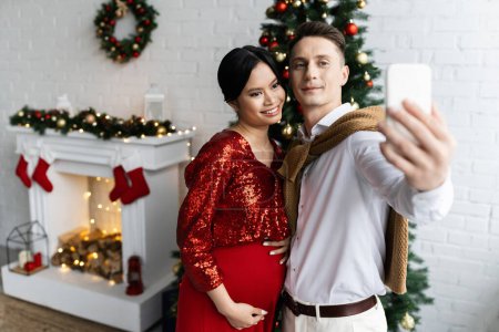 junger Mann macht Selfie mit glücklicher und schwangerer asiatischer Frau in der Nähe von Weihnachtsbaum und dekoriertem Kamin
