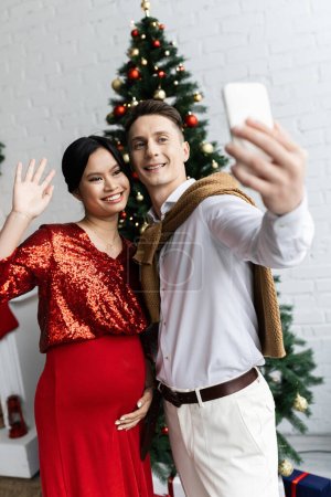 glücklicher Mann mit Handy in der Nähe schwangere asiatische Frau winkt bei Videoanruf am Weihnachtstag