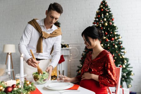 Schwangere Asiatin lächelt beim romantischen Weihnachtsessen zu Hause neben Mann mit frischem Gemüsesalat