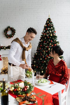 happy man holding bowl of vegetable salad near pregnant asian woman during Christmas supper at home