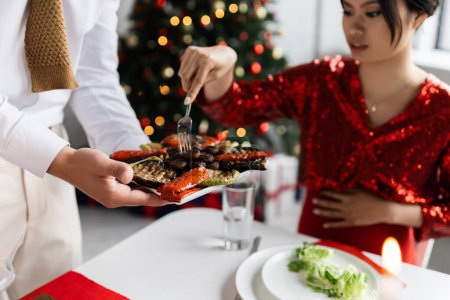 man holding grilled vegetables near pregnant asian wife with fork during romantic Christmas supper on blurred background