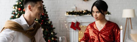 lächelnder Mann, der auf eine seriöse asiatische Frau in roter und glänzender Bluse schaut, während sie zu Hause Weihnachten feiert, Banner