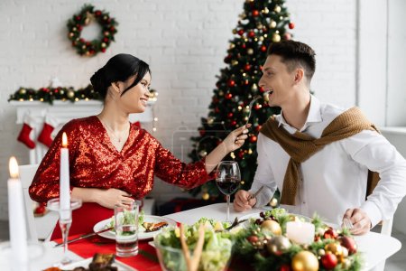 pregnant and cheerful asian woman feeding husband while having fun at romantic Christmas supper
