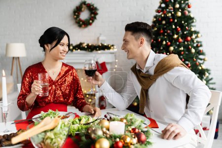 pregnant asian woman with husband holding glasses with drinks during romantic Christmas supper in living room