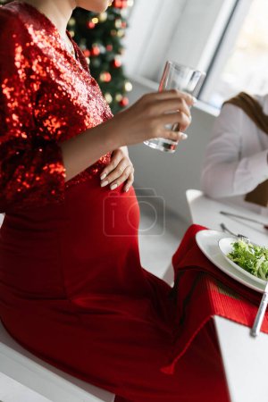 geschnittene Ansicht einer schwangeren Frau, die beim Weihnachtsessen mit ihrem Mann ein Glas Wasser in der Nähe von frischem Salat hält
