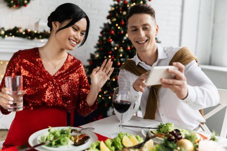 smiling man holding smartphone near pregnant asian wife waving hand during video call during festive supper