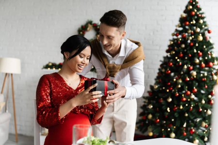 glücklich und schwanger asiatische Frau nimmt Weihnachtsgeschenk von jungen Mann