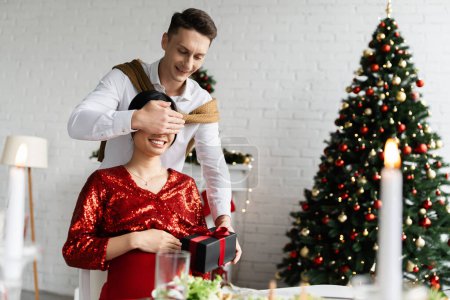 cheerful man covering eyes of pregnant asian wife while holding Christmas gift