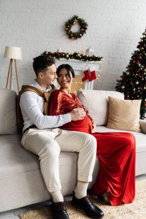 young man touching tummy of pregnant asian wife in festive clothing on cozy sofa near blurred Christmas decor