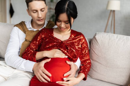 Photo for Young man embracing belly of pregnant asian wife wearing red and shiny clothes - Royalty Free Image