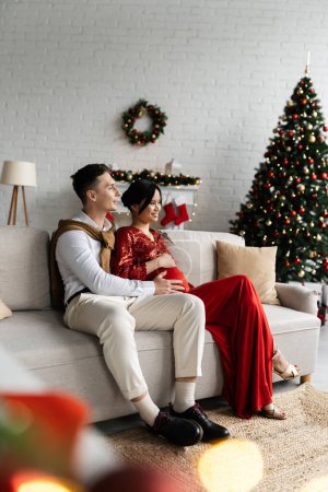 Foto de Feliz hombre con embarazada asiático esposa en festivo traje sentado en acogedor sofá en moderno salón con decoración de Navidad - Imagen libre de derechos