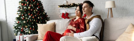 young man and pregnant asian woman in festive clothing on cozy sofa in living room with Christmas tree, banner