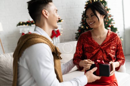 Téléchargez les photos : Femme asiatique enceinte souriant au mari présentant cadeau de Noël sur le canapé à la maison - en image libre de droit